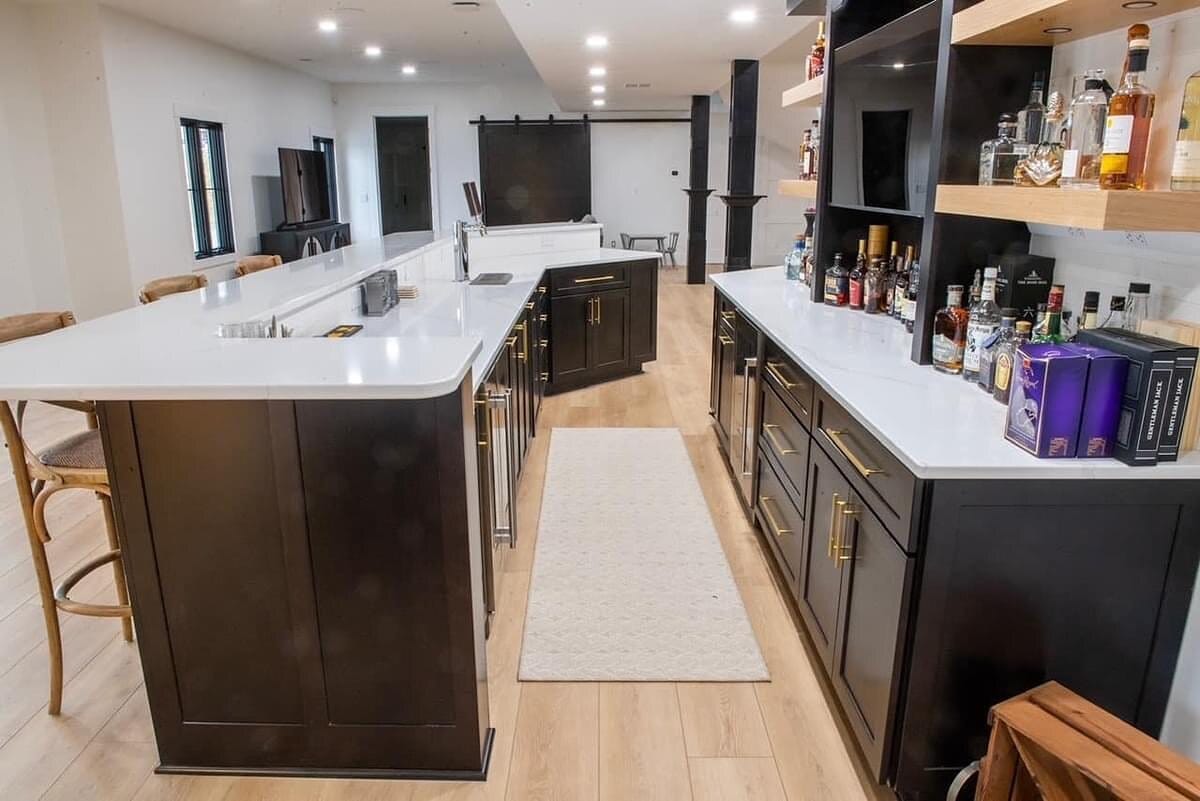 Wide view of a basement bar and lounge with dark cabinetry, an L-shaped quartz counter, open liquor shelving, and rattan stools.