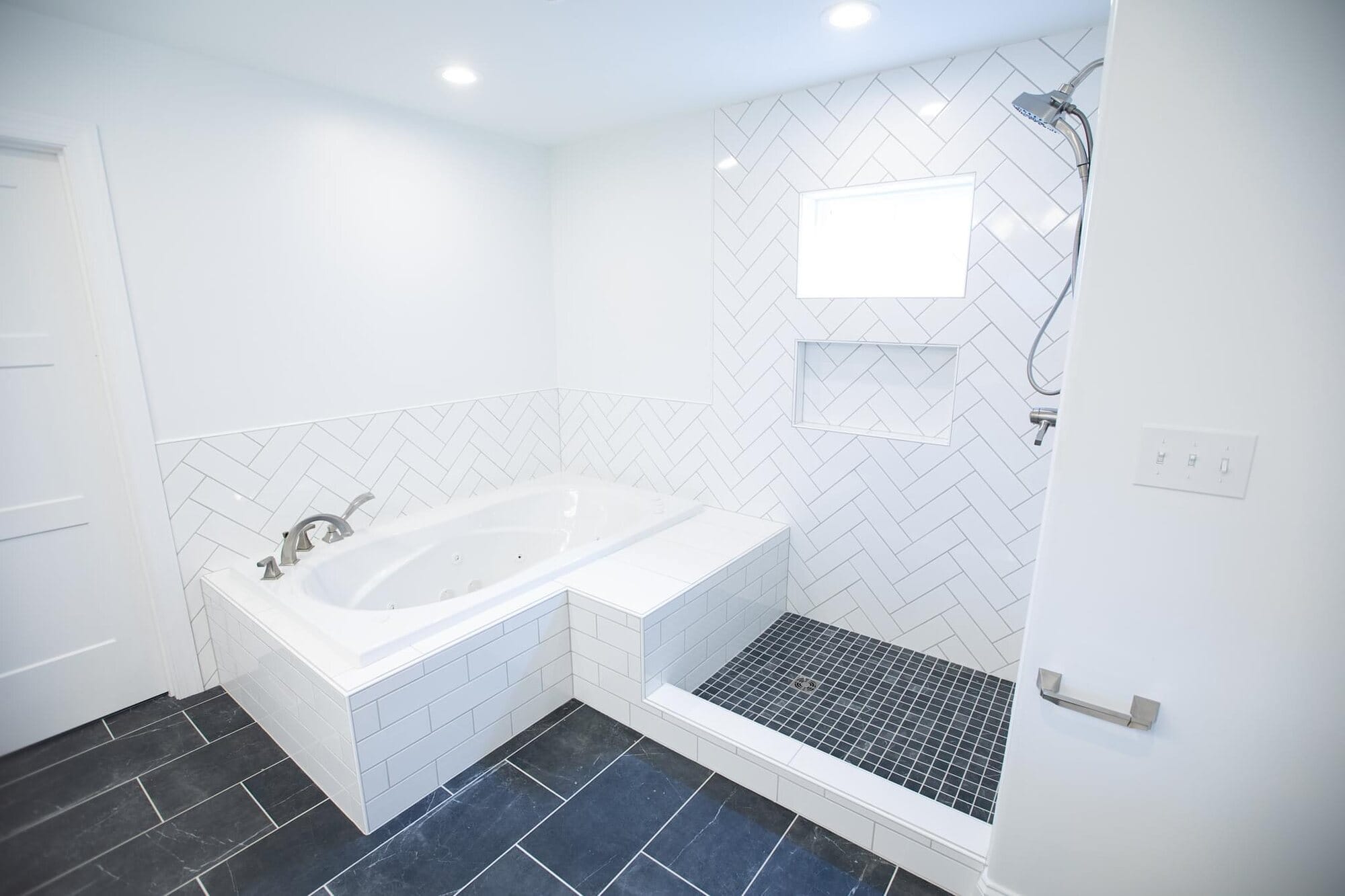 Bathroom with a corner drop-in soaking tub, adjacent walk-in shower with herringbone tile, and dark slate-look floor tile.