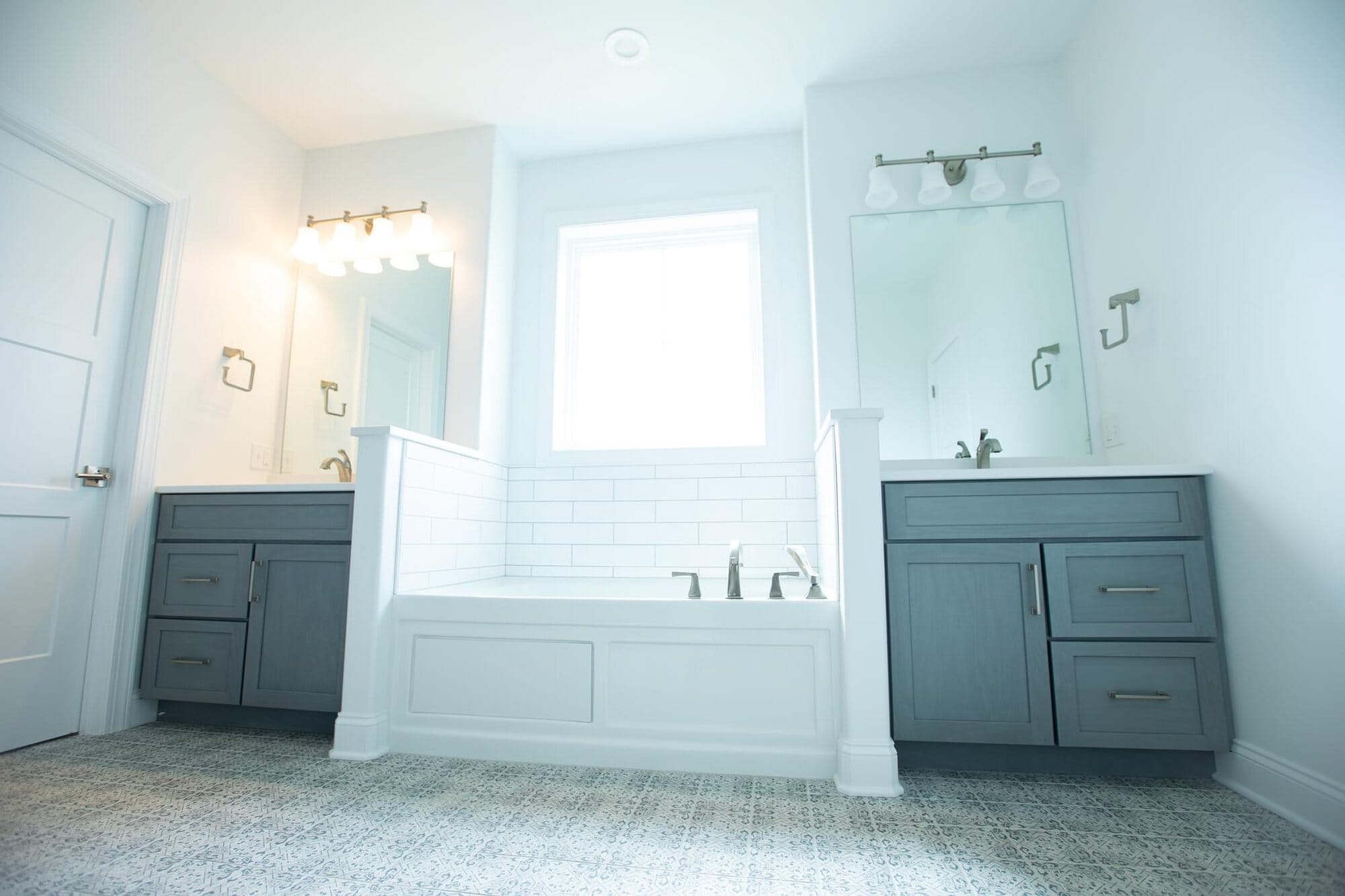 Bathroom with a drop-in soaking tub between two separate gray vanities, framed mirrors, and brushed gold sconces.