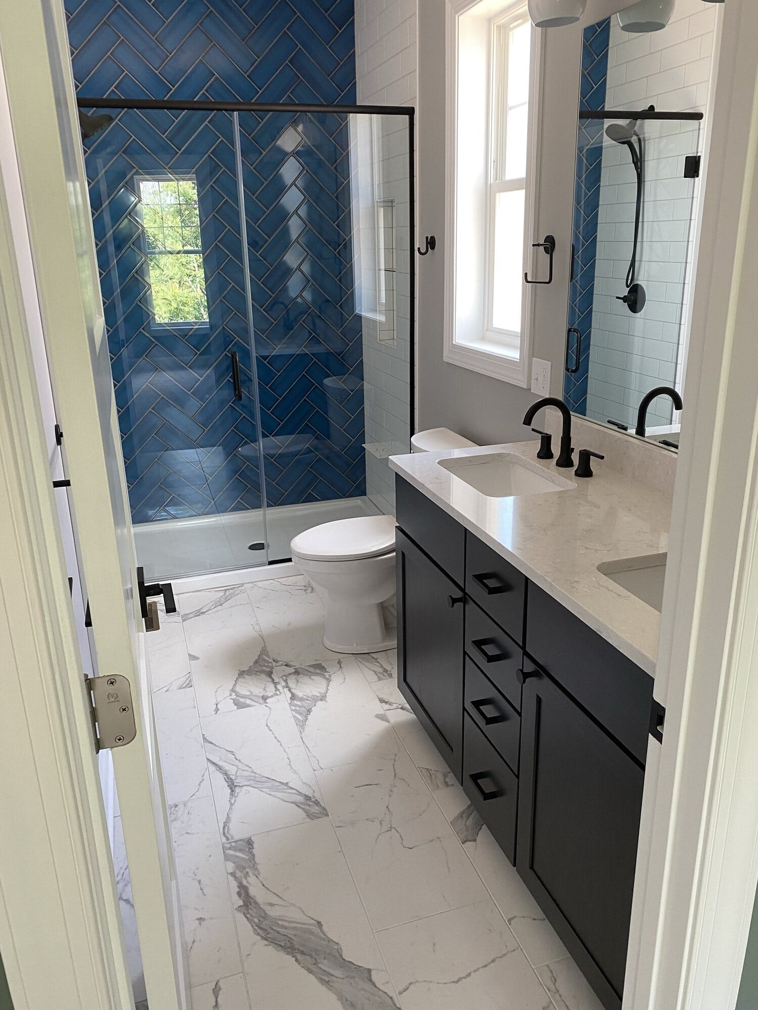 Bathroom remodel with a navy chevron tile shower, matte black fixtures, dark double vanity, and marble-look porcelain flooring.