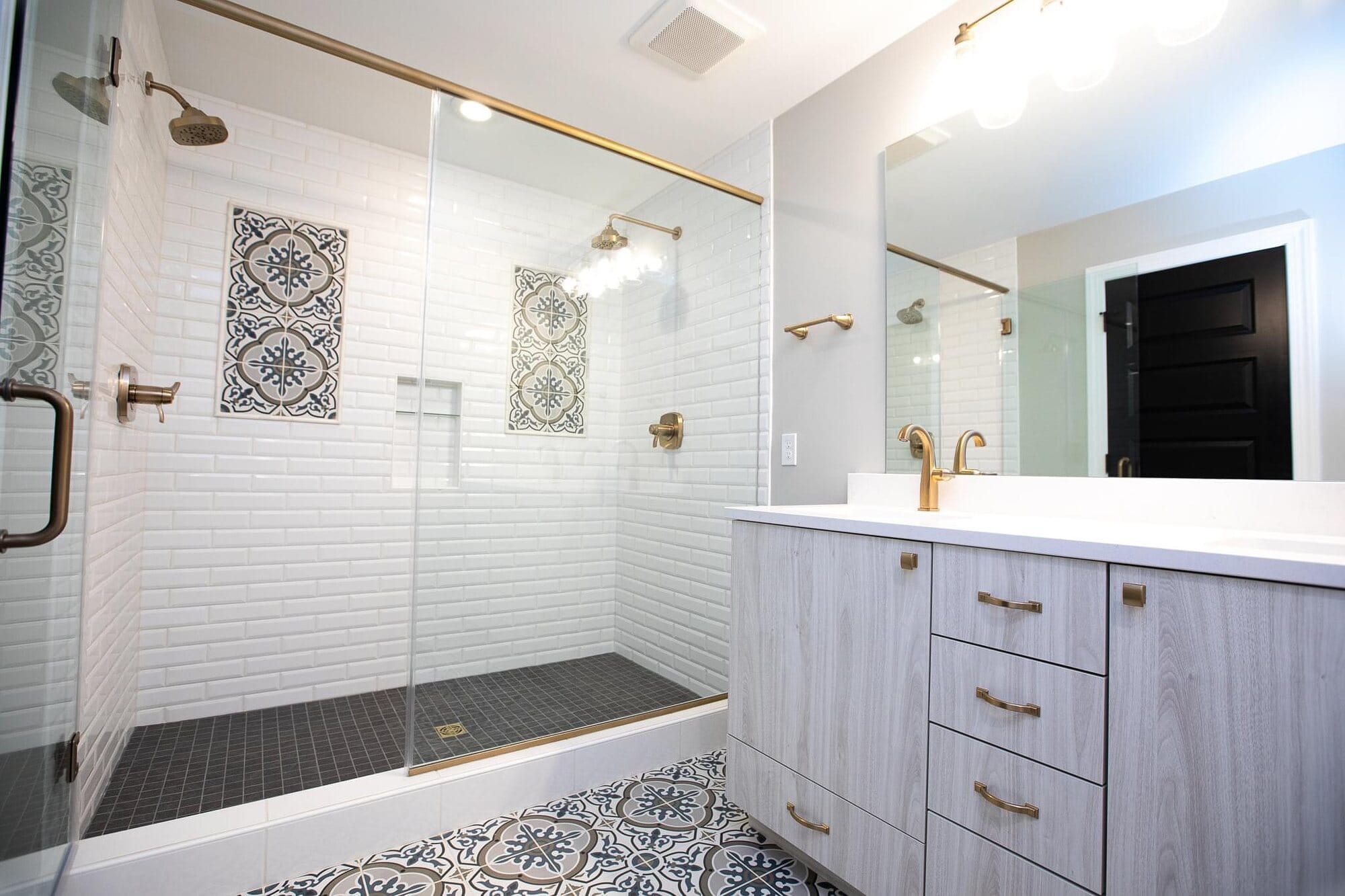 Bathroom featuring a white subway tile shower with decorative accent inserts, brushed gold fixtures, light wood vanity, and patterned floor tile.
