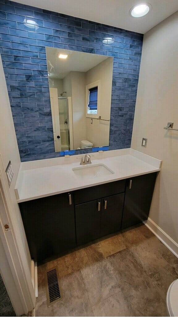 Bathroom vanity with a dark slab-front cabinet, white quartz top, and a blue stacked stone accent wall behind a frameless mirror.