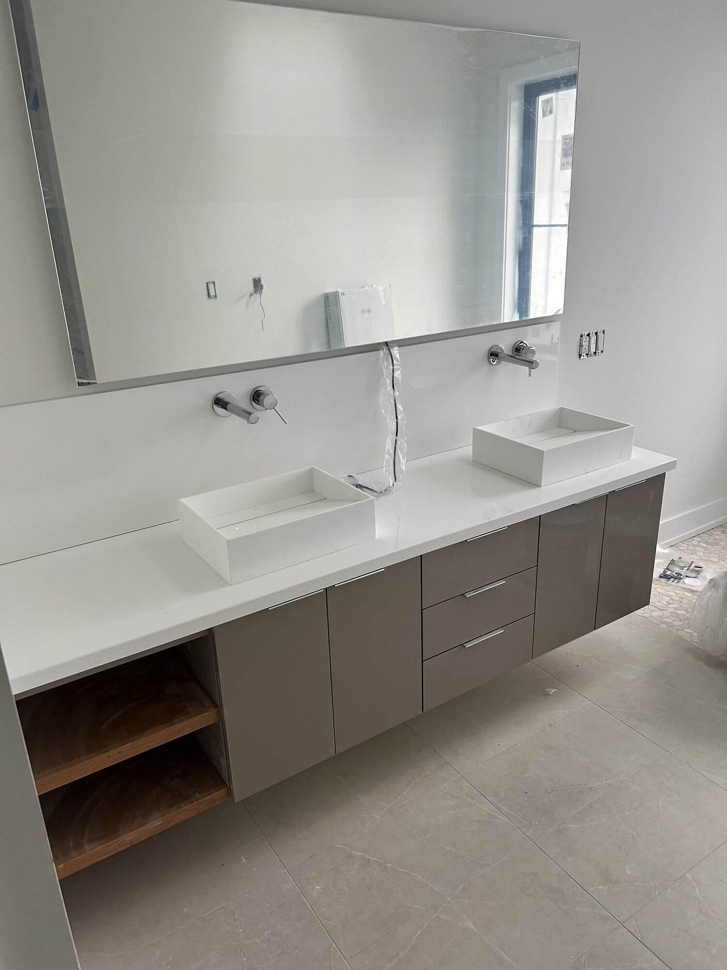Modern floating double vanity in taupe slab cabinetry with rectangular vessel sinks, wall-mount faucets, and open wood shelving.
