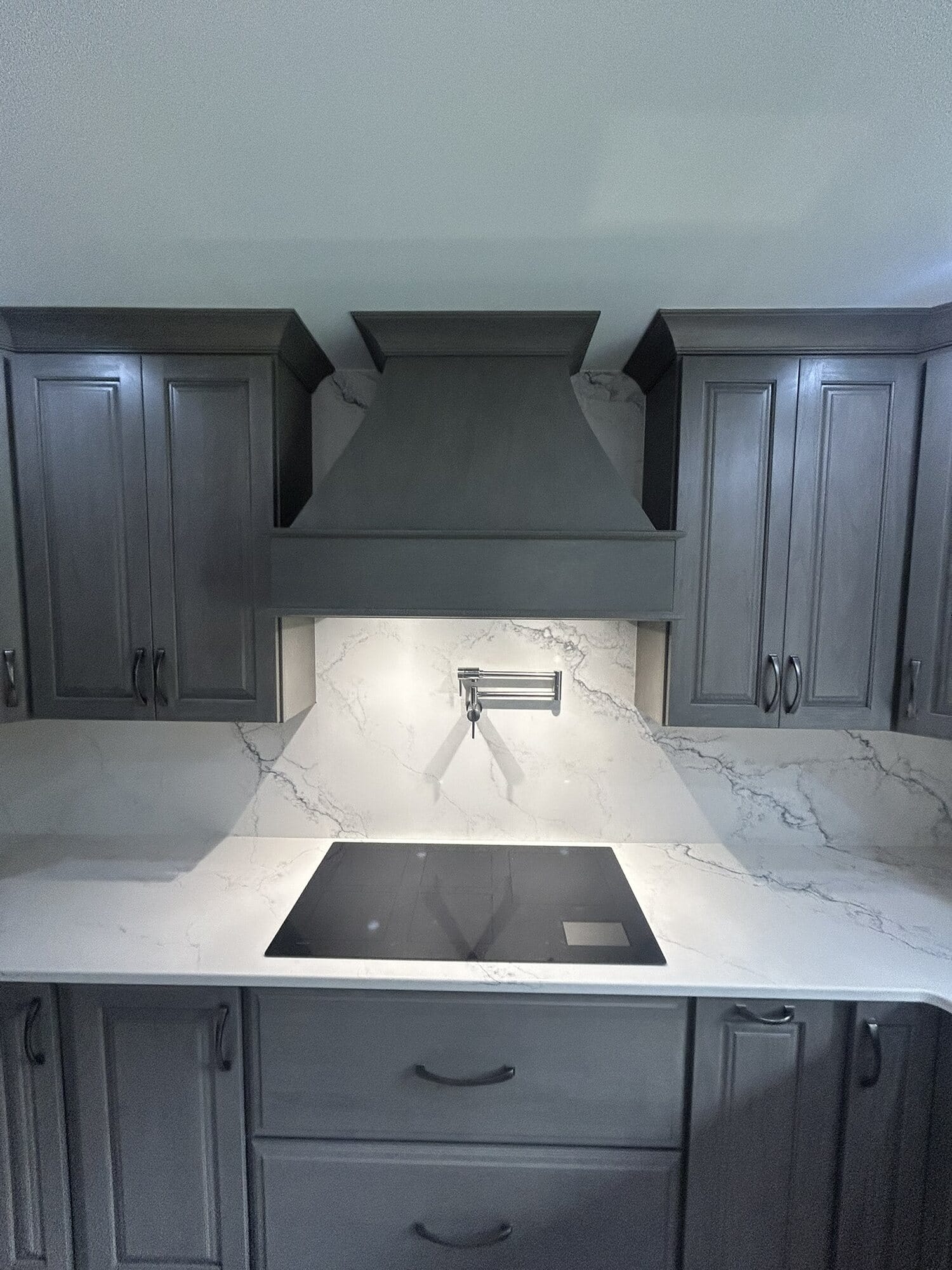 Kitchen range wall with a custom gray stained hood, induction cooktop, and full-height marble-look quartz backsplash.