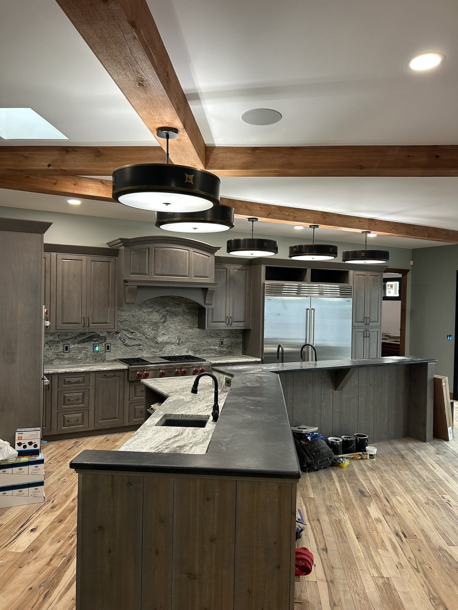 Kitchen with gray stained cabinetry, dark honed countertops, a raised bar island, wood beams, and drum pendant lighting.