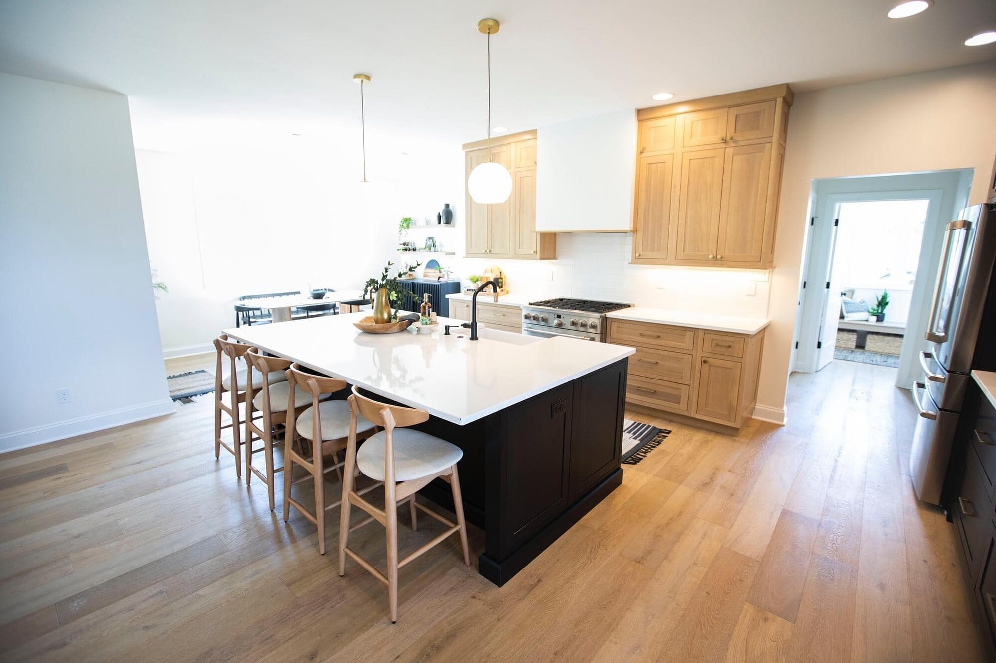 Open kitchen with natural wood upper cabinetry, a dark painted island with quartz top, counter stools, and white oak flooring.