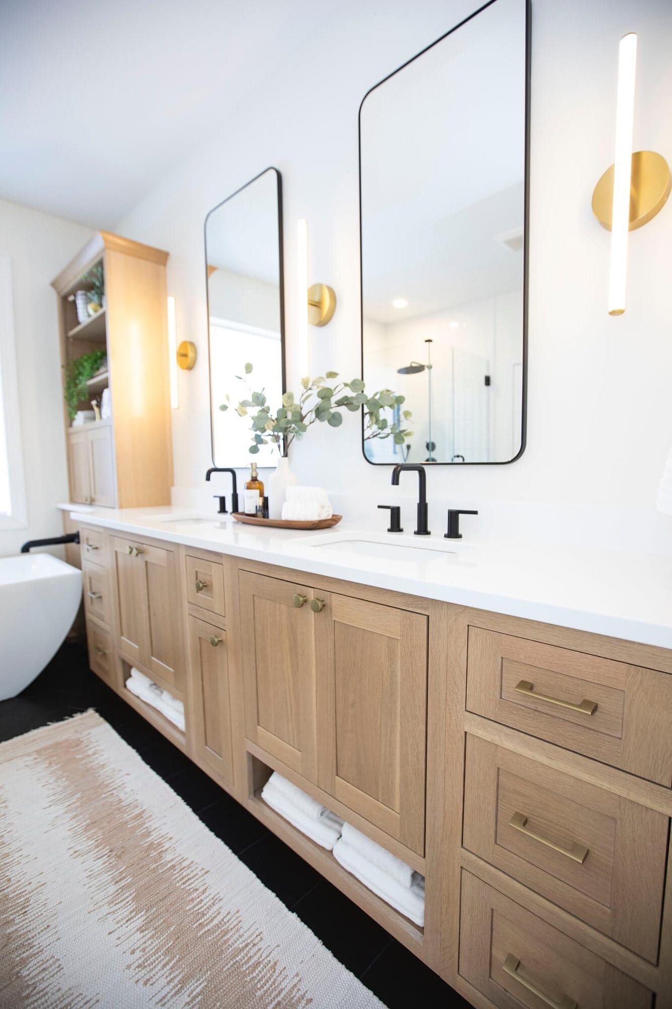 Master bathroom with a light oak double vanity, quartz top, backlit sconces, arched mirrors, and a freestanding tub.