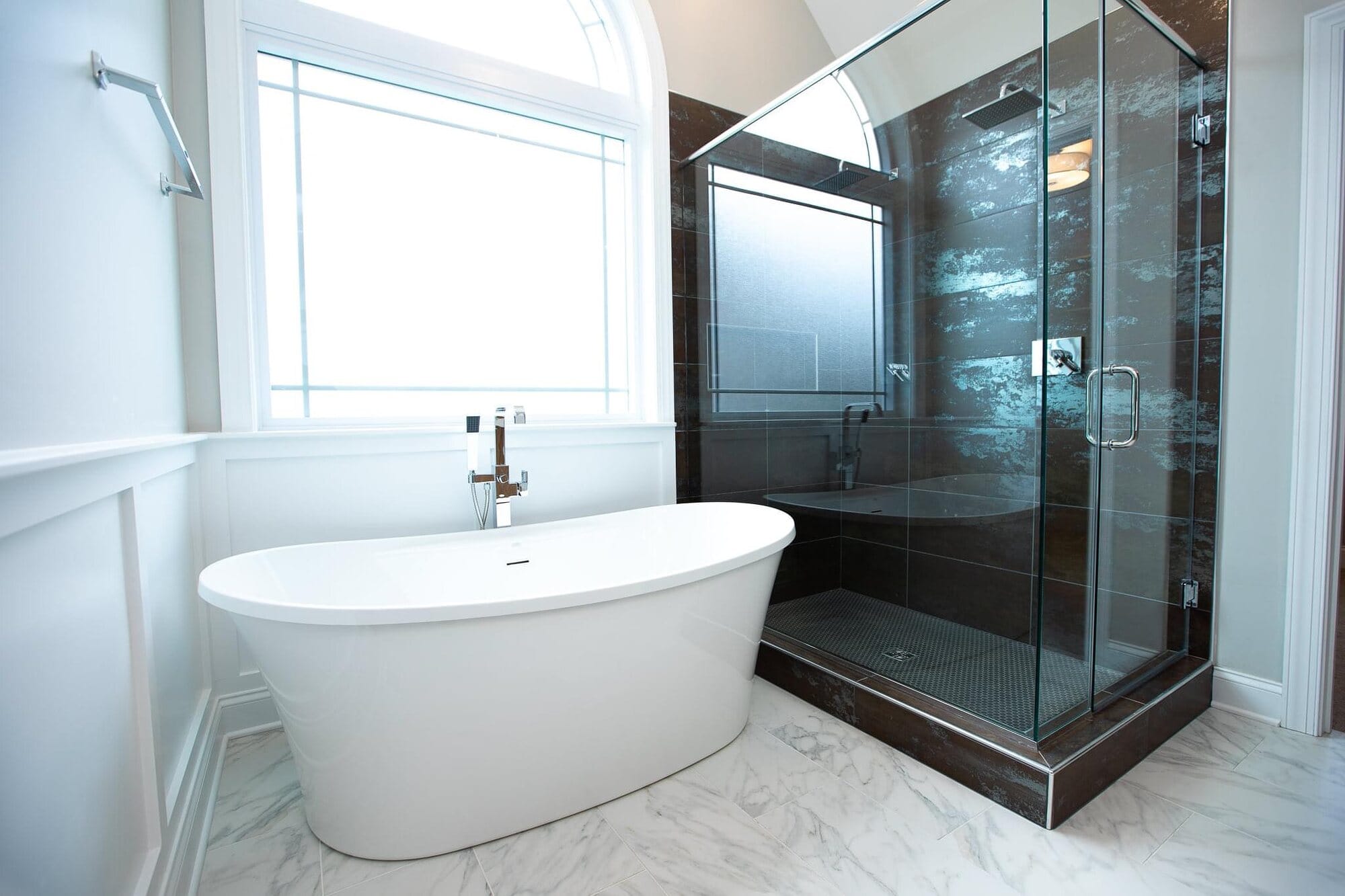 Master bathroom with a freestanding tub beneath an arched window, smoked glass shower enclosure, and marble-look tile floor.