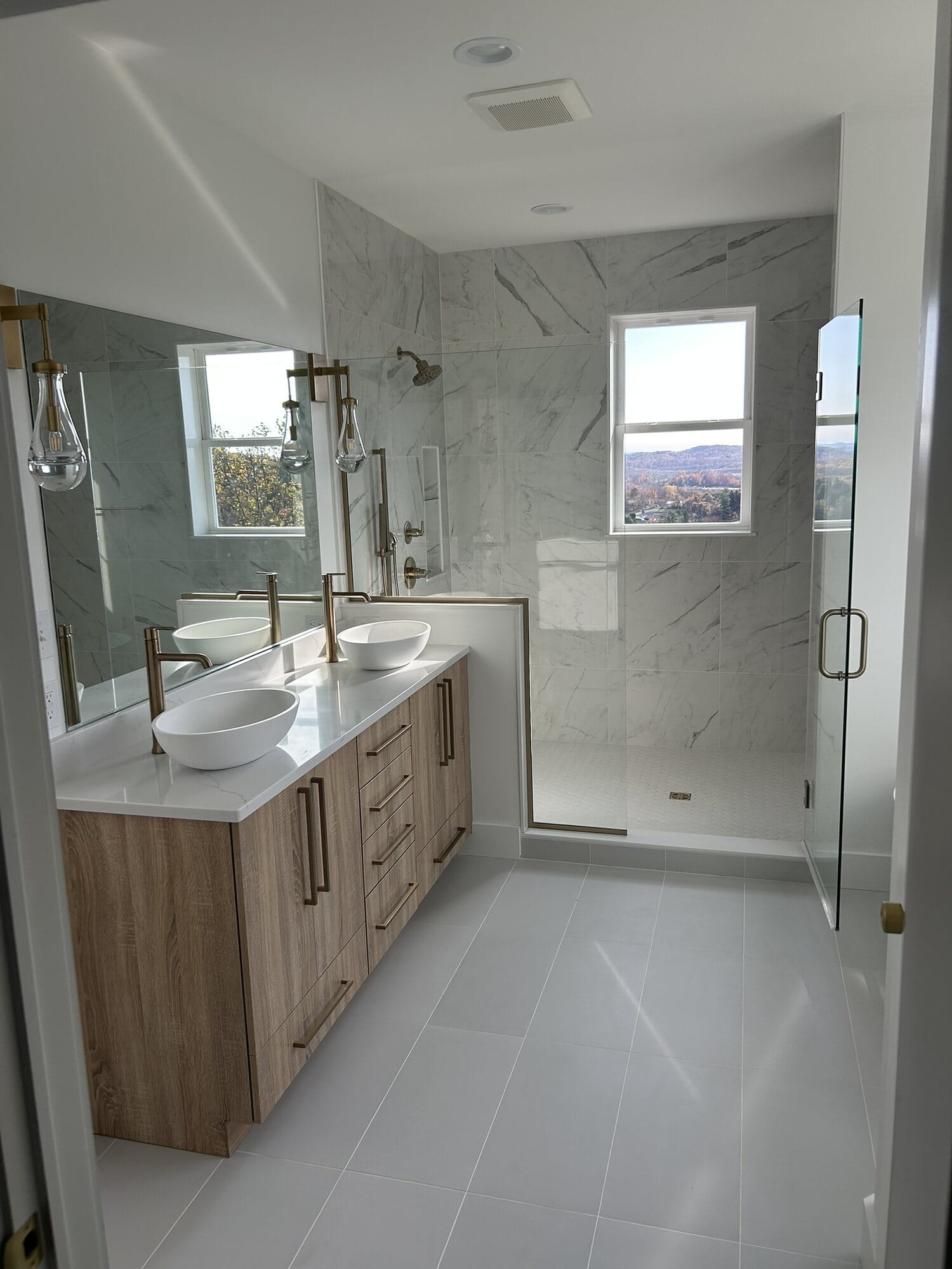 Master bathroom with an oak double vanity, vessel sinks, frameless marble-tiled shower, and large-format gray floor tile.