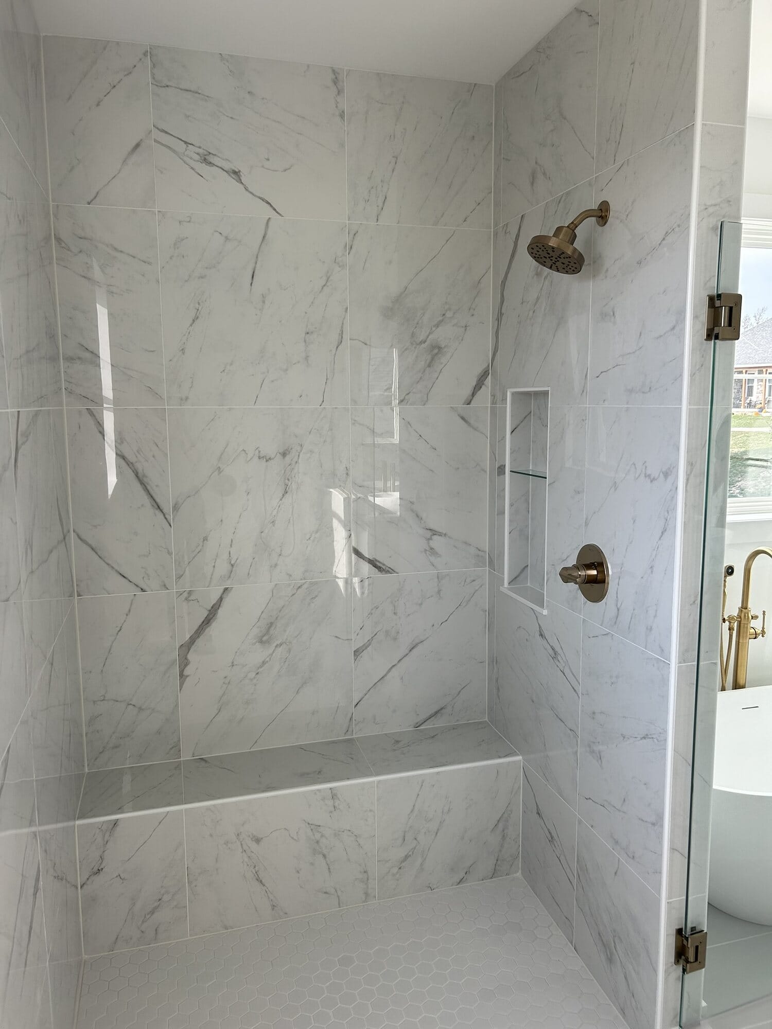 Tub and shower surround in marble-look panels with brushed gold fixtures, a recessed niche, and glass panel.