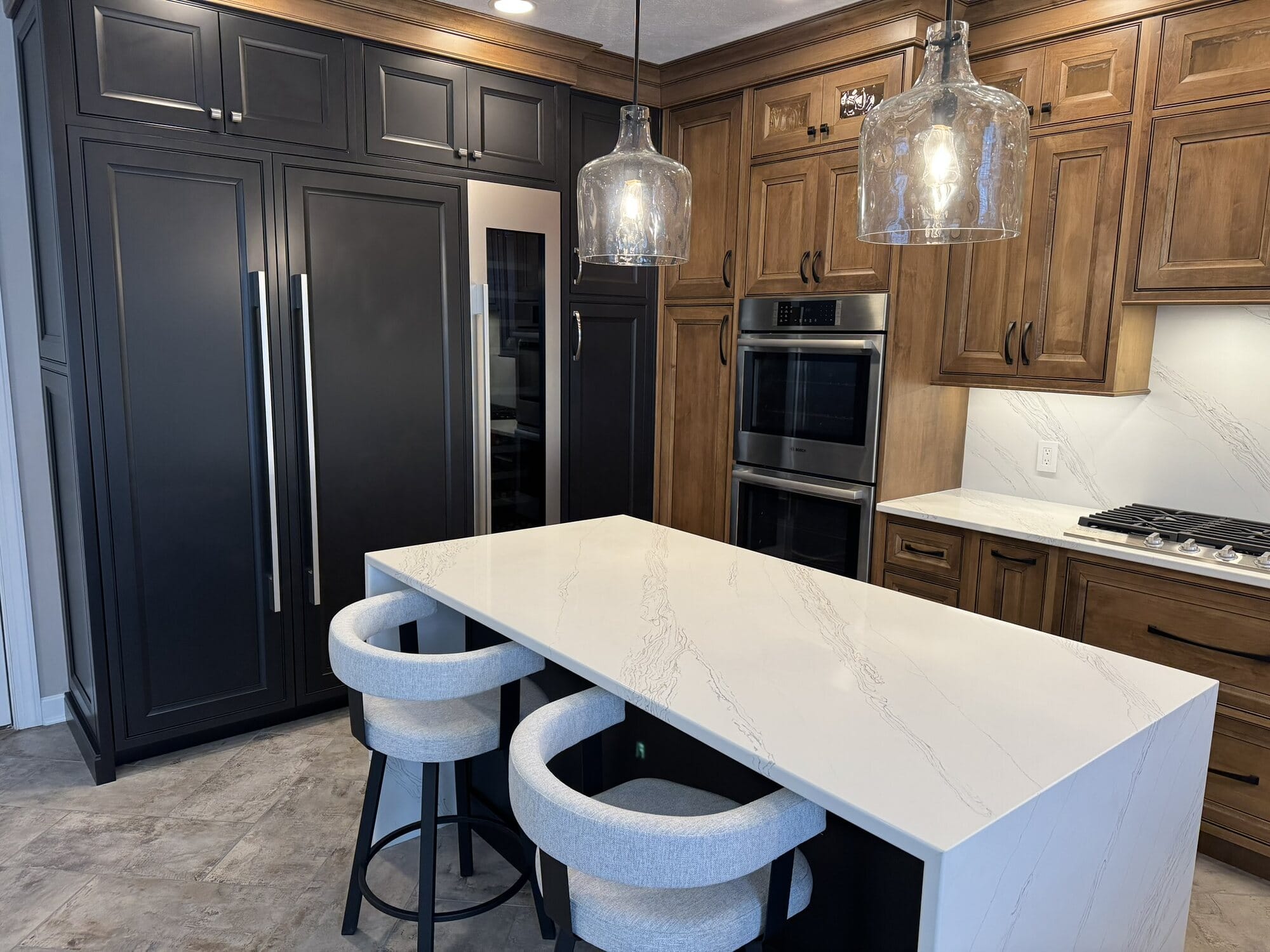 Two-tone kitchen pairing black painted cabinetry with stained wood, topped by a waterfall-edge quartz island with upholstered stools.
