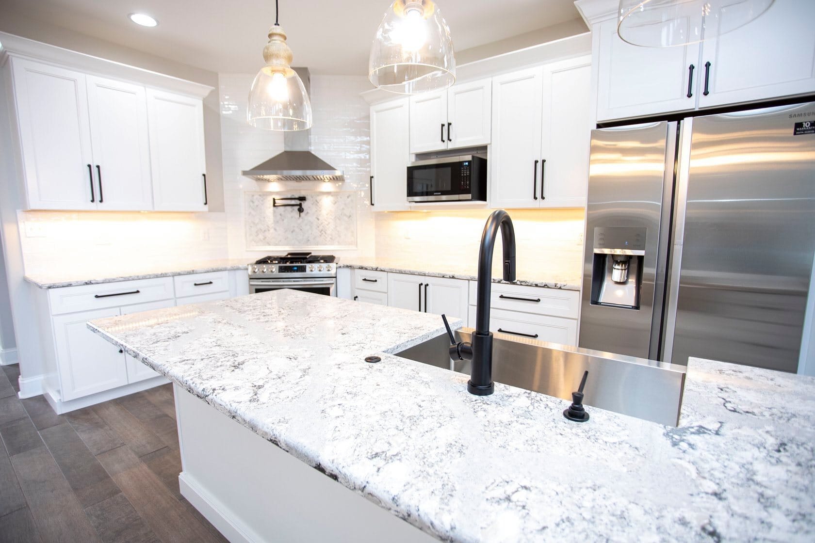 White kitchen with a granite-topped island, matte black gooseneck faucet, glass pendant lights, and stainless steel appliances.