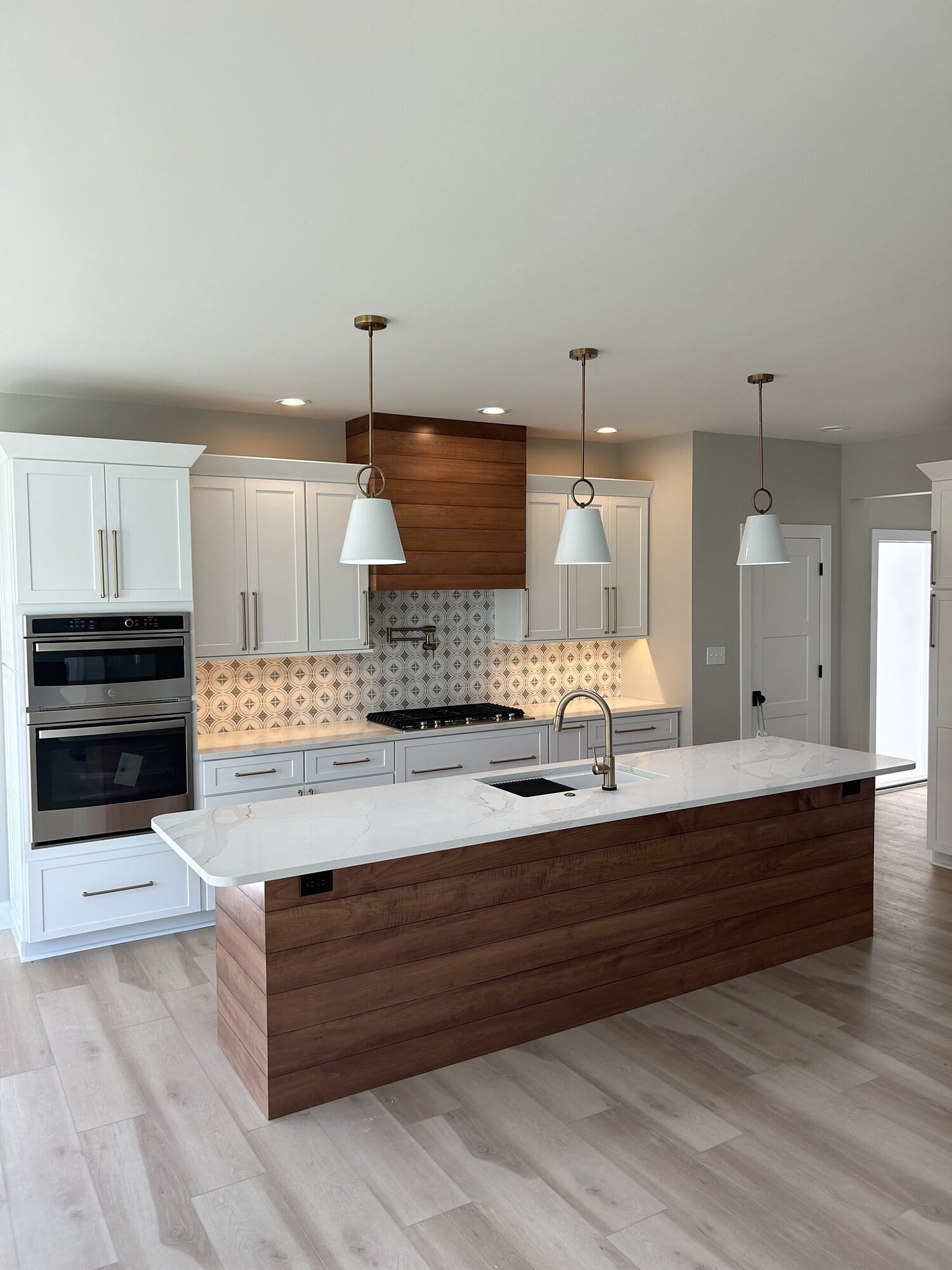 White kitchen with a walnut-front island, custom wood vent hood, patterned tile backsplash, and wall ovens.