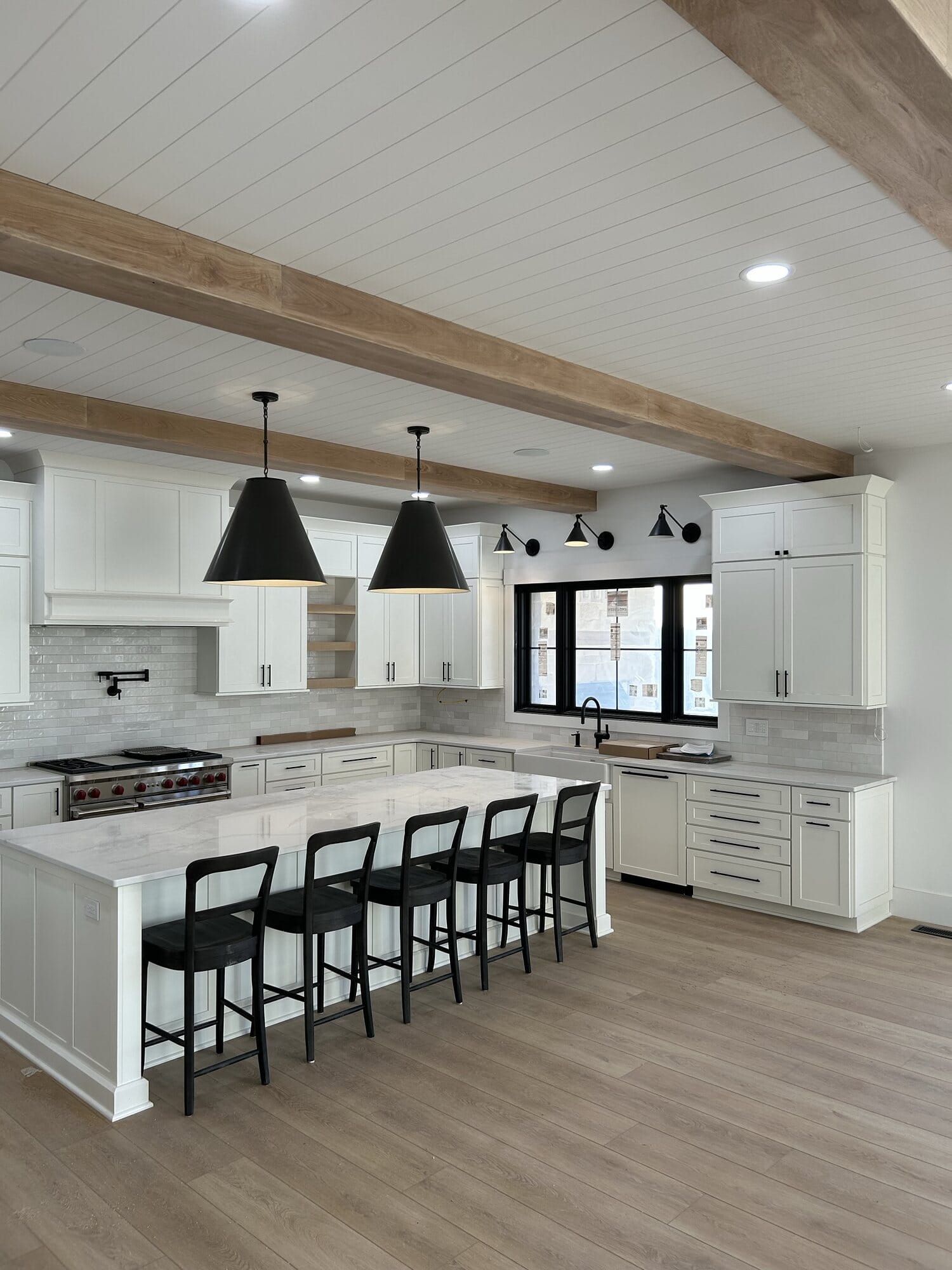 White kitchen with a shiplap and wood beam ceiling, extended island with black stools, black pendants, and pro-style range.