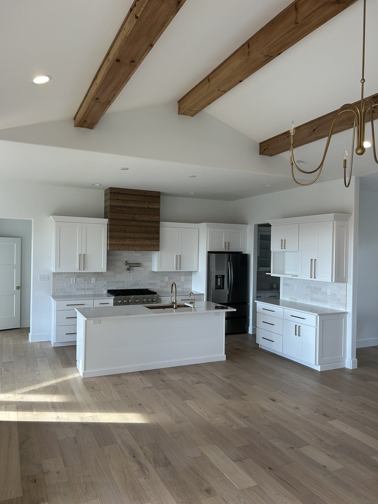 White kitchen with exposed wood ceiling beams, a stained wood vent hood, quartz island, and black panel-ready refrigerator.